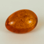 Polished oval amber gemstone with warm golden-orange tones and visible natural inclusions, photographed on a neutral background under soft lighting.